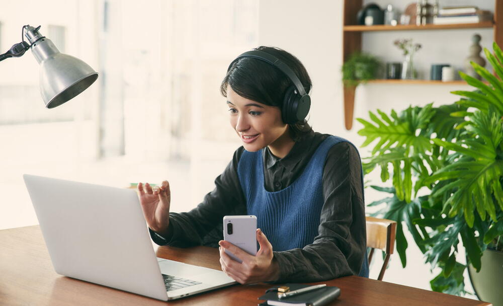 Imagine cu o femeie la birou, purtand o pereche de casti Sony WH-CH720 negre si folosind un laptop si un telefon mobil