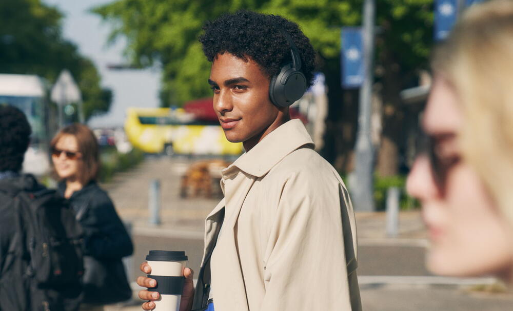 Imagine cu un barbat in oras, purtand o pereche de casti Sony WH-CH720 negre si tinand o ceasca de cafea