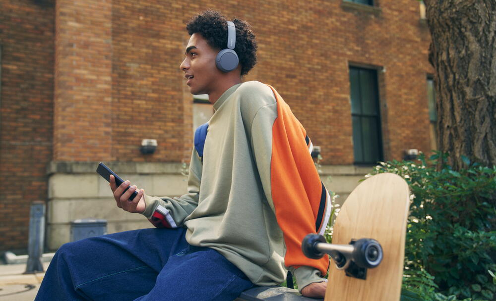 Imagine cu un barbat pe un perete, cu castile Sony WH-CH520 si tinand un telefon mobil, cu un skateboard in prim-plan